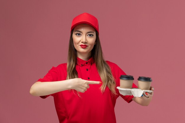 Front view female courier in red uniform holding brown delivery coffee cups on light-pink background service delivery uniform worker job female lady company