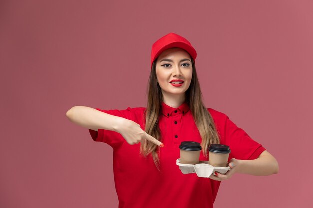 Front view female courier in red uniform holding brown delivery coffee cups on pink background service delivery uniform job