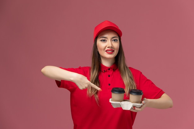 ピンクの背景に茶色の配達コーヒーカップを保持している赤い制服の正面図女性宅配便サービス配達制服の仕事