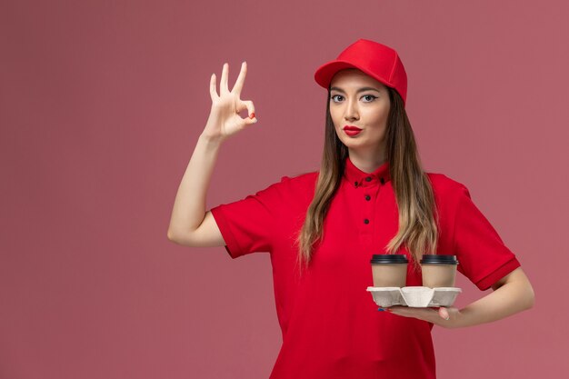 Front view female courier in red uniform holding brown delivery coffee cups posing on pink background service job delivery uniform