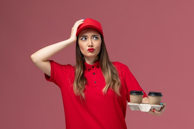 Front view female courier in red uniform holding brown delivery coffee cups and thinking on light-pink background service delivery uniform worker job female company