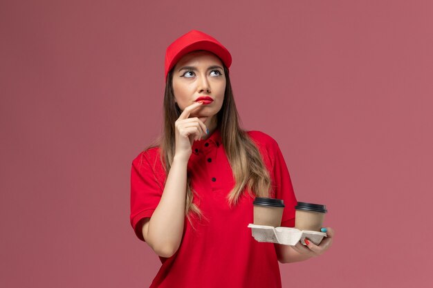 Front view female courier in red uniform holding brown delivery coffee cups and thinking on pink background service delivery uniform worker job female company