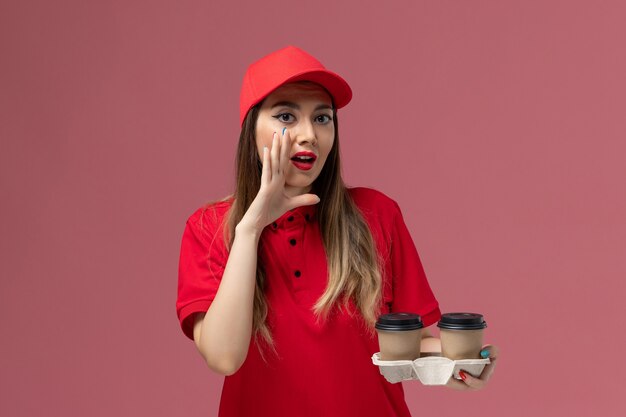 Front view female courier in red uniform holding brown delivery coffee cups whispering on light-pink background service delivery uniform worker job female company