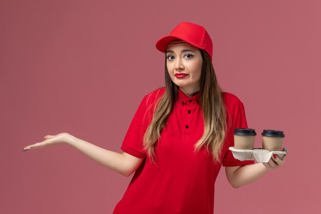 ピンクの背景のサービス配達制服会社で混乱した表情で茶色の配達コーヒーカップを保持している赤い制服の正面図女性宅配便