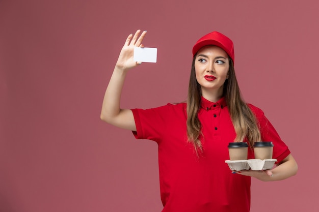 Вид спереди женщина-курьер в красной униформе держит доставку кофейных чашек и открытку на розовом фоне.