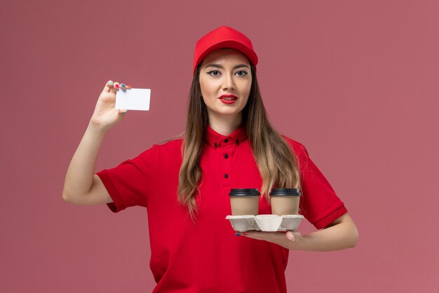 Front view female courier in red uniform holding delivery coffee cups and card on pink background service job delivery uniform