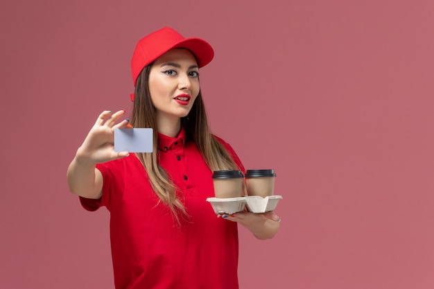 Front view female courier in red uniform holding delivery coffee cups and card posing on pink background service job delivery uniform