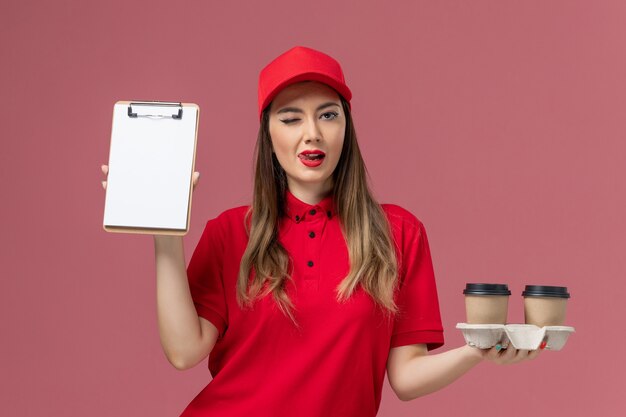 淡いピンクの背景に配達コーヒーカップとメモ帳を保持している赤い制服の正面図女性宅配便サービス配達仕事労働者の制服