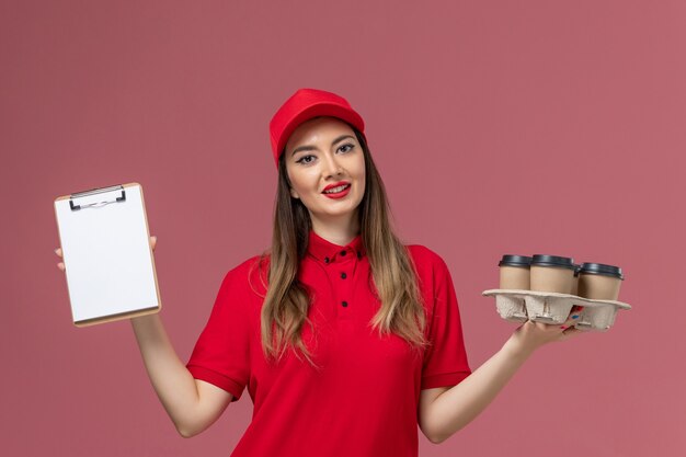 ピンクの背景に配達コーヒーカップメモ帳を保持している赤い制服の正面図女性宅配便サービス配達会社の仕事の制服