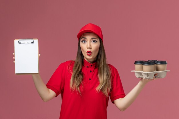 ピンクの背景のサービス配達会社の制服に配達コーヒーカップとメモ帳を保持している赤い制服の正面図女性宅配便