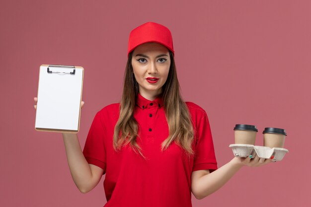 ピンクの背景に配達コーヒーカップとメモ帳を保持している赤い制服の正面図女性宅配便サービス配達労働者の制服