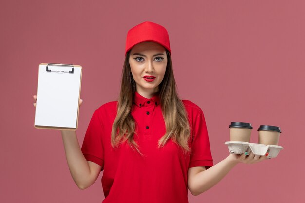 Front view female courier in red uniform holding delivery coffee cups and notepad on pink background service delivery job worker uniform