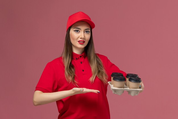 Вид спереди женщина-курьер в красной форме, держащая доставку кофейных чашек на розовом фоне, форма доставки службы, рабочий