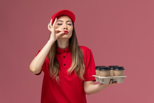 Front view female courier in red uniform holding delivery coffee cups showing tasty sign on pink background service delivery uniform job worker