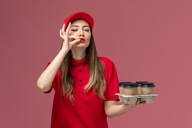 Front view female courier in red uniform holding delivery coffee cups showing tasty sign on pink background service delivery uniform job worker