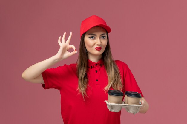 ピンクの背景に笑顔の配達コーヒーカップを保持している赤い制服の正面図女性宅配便労働者の仕事サービス配達制服