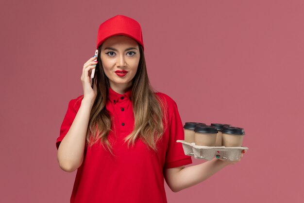 Вид спереди женщина-курьер в красной форме, держащая кофейные чашки доставки и разговаривающая по телефону на розовом столе, служба доставки униформы