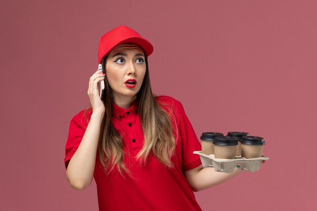 Front view female courier in red uniform holding delivery coffee cups and talking on the phone on pink desk service delivery uniform