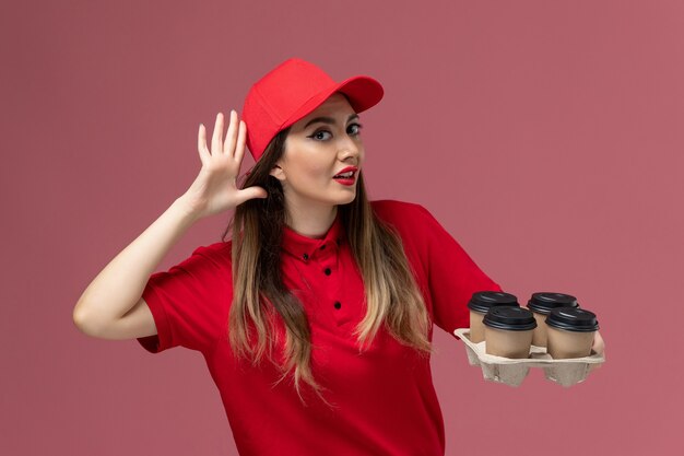 Вид спереди женщина-курьер в красной форме, держащая кофейные чашки с доставкой, пытается выслушать на розовом фоне работник службы доставки униформы