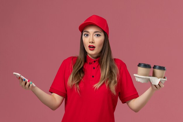 배달 커피 컵을 들고 밝은 분홍색 배경 작업자 작업 서비스 배달 유니폼에 전화를 사용하는 빨간 제복을 입은 전면보기 여성 택배