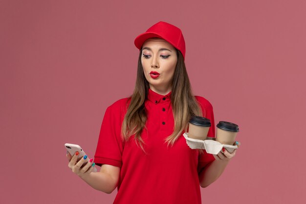 배달 커피 컵을 들고 분홍색 배경 작업자 작업 서비스 배달 유니폼에 전화를 사용하는 빨간색 제복을 입은 전면보기 여성 택배