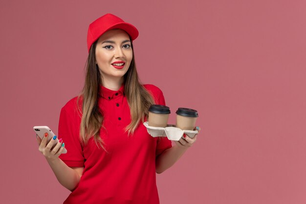 Front view female courier in red uniform holding delivery coffee cups and using phone on the pink background worker job service delivery uniform