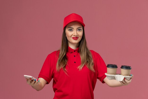 Front view female courier in red uniform holding delivery coffee cups and using phone smiling on pink background worker job service delivery uniform