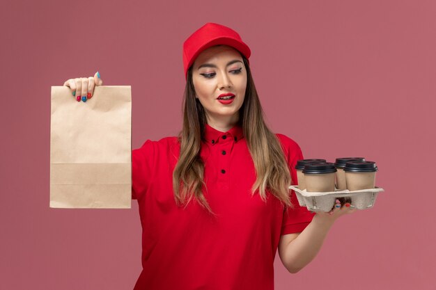 ピンクの背景に食品パッケージと配達コーヒーカップを保持している赤い制服の正面図女性宅配便