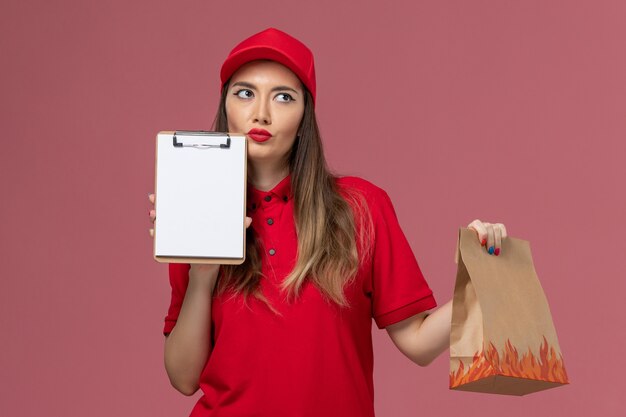 Front view female courier in red uniform holding notepad and food package thinking on the pink background service job delivery uniform company