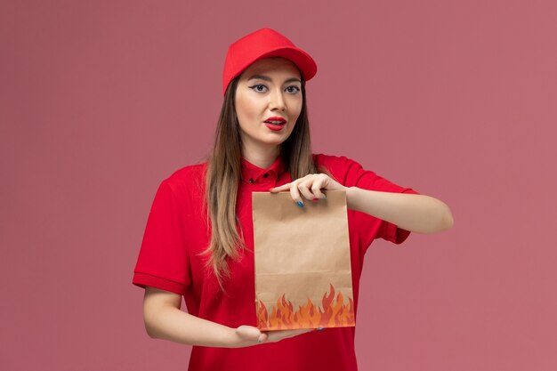 Front view female courier in red uniform holding paper food package on light-pink background service delivery uniform company job