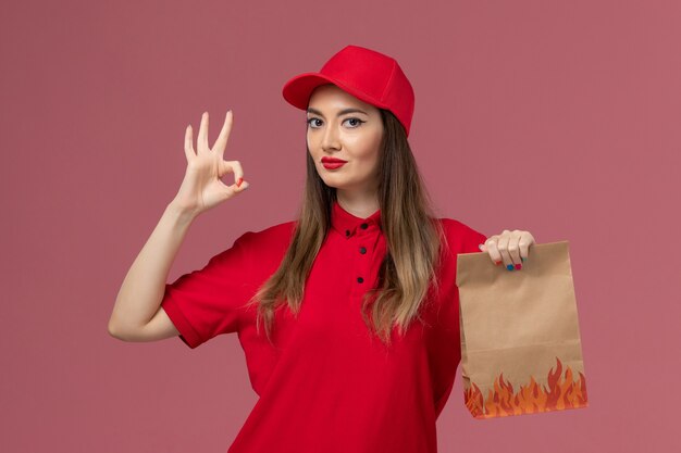 Front view female courier in red uniform holding paper food package on the light pink background service job delivery uniform company