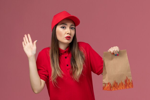 Front view female courier in red uniform holding paper food package on the light-pink background worker service job delivery uniform company