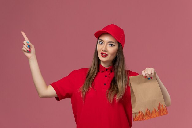 Front view female courier in red uniform holding paper food package on the pink background service delivery uniform company