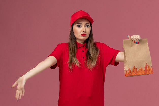ピンクの背景に紙の食品パッケージを保持している赤い制服の正面図の女性の宅配便
