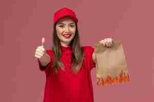 Foto gratuita corriere femminile di vista frontale in uniforme rossa che tiene il pacchetto alimentare di carta che sorride sull'azienda dell'uniforme di consegna di lavoro di servizio del fondo rosa