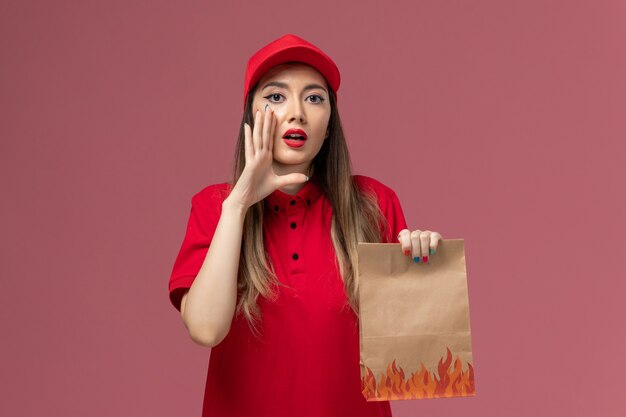 Front view female courier in red uniform holding paper food package whispering on the pink background worker service delivery uniform company job