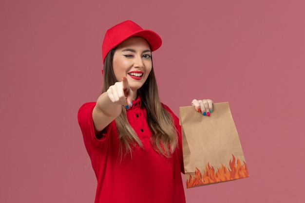 Front view female courier in red uniform holding paper food package winking on pink background service job delivery uniform company