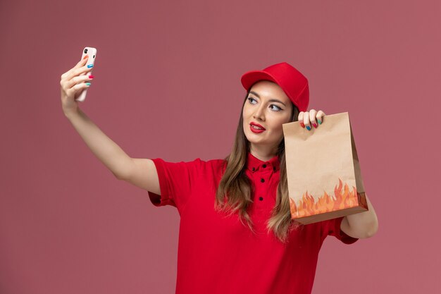 Front view female courier in red uniform holding phone and food package taking photo on the pink background service delivery uniform job worker