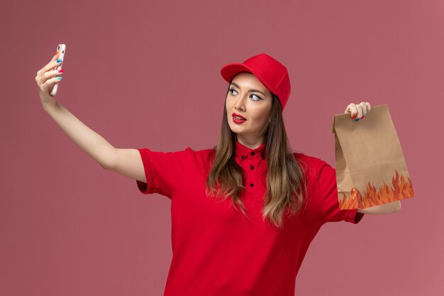 Front view female courier in red uniform holding phone and food package taking selfie on pink background service delivery uniform
