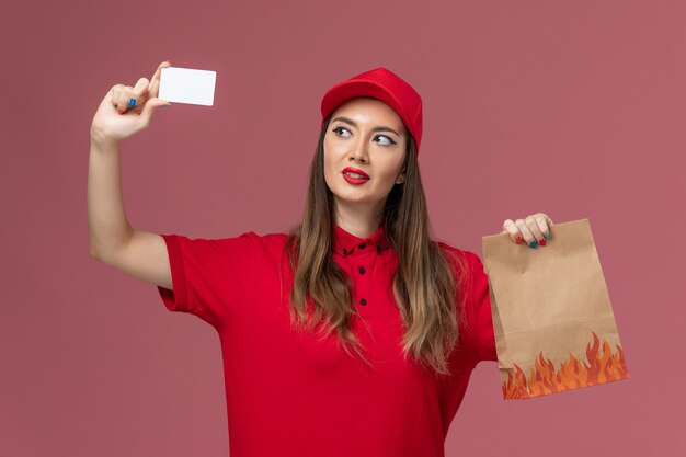 Вид спереди женщина-курьер в красной форме, держащая белую карточку с продуктовым пакетом на светло-розовом фоне, служба доставки работы, униформа компании