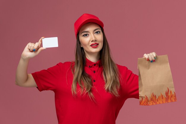 Front view female courier in red uniform holding white card and food package on the pink background service job delivery uniform company