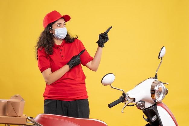 Front view female courier in red uniform and mask on yellow background covid- job delivery uniform worker pandemic