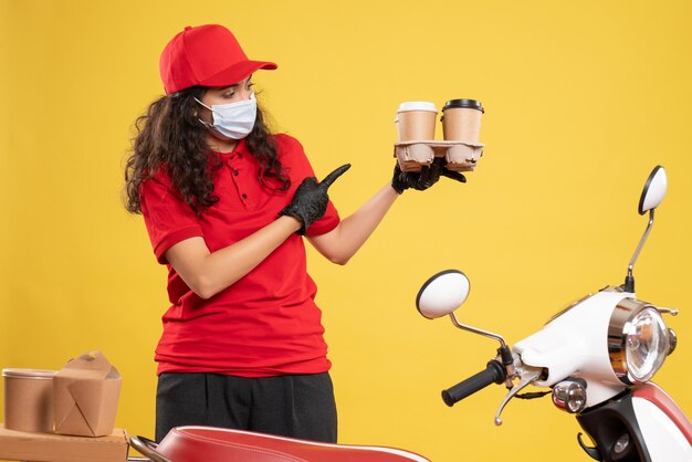 Front view female courier in red uniform with coffee cups on yellow background delivery covid- pandemic job uniform service virus