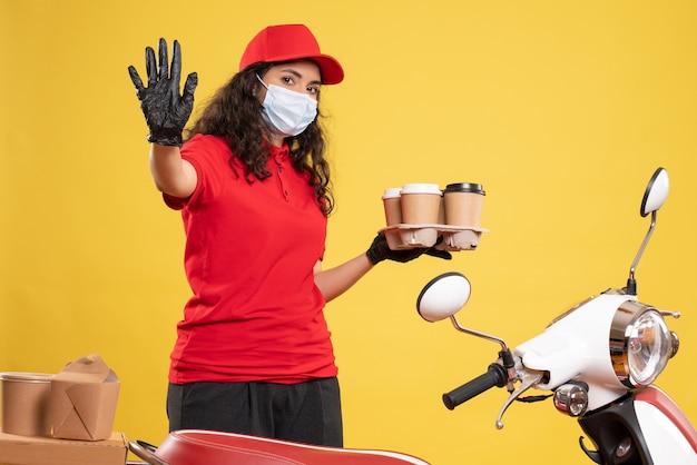 Front view female courier in red uniform with coffee cups on yellow background worker covid- pandemic job uniform service virus