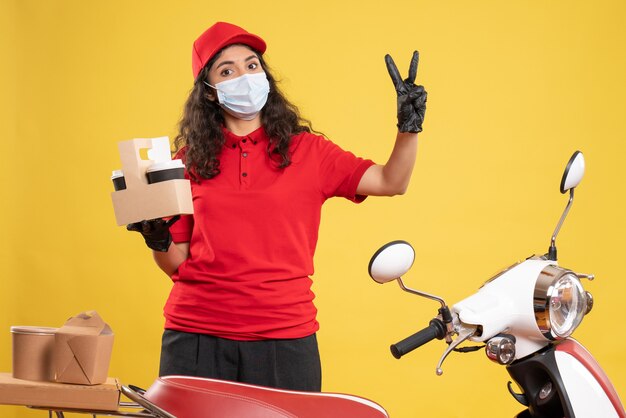 Front view female courier in red uniform with coffee cups on yellow background worker delivery covid- pandemic uniform service virus