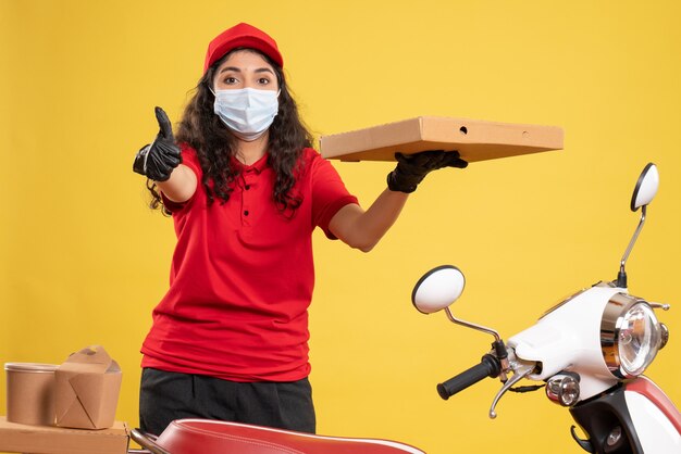 Front view female courier in red uniform with pizza box on yellow desk service worker covid- pandemic virus job delivery