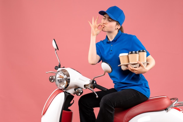 Corriere femminile di vista frontale che si siede sulla bici con le tazze di caffè sul servizio alimentare dell'operaio di consegna uniforme di colore di lavoro rosa