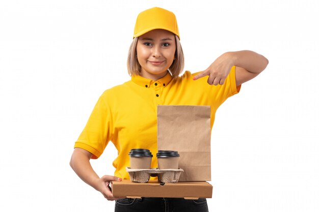 A front view female courier in yellow cap yellow shirt smiling and holding coffee boxes posing on white