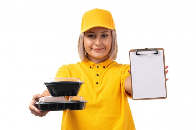 A front view female courier in yellow shirt yellow cap and black jeans holding bowls with food and notepad smiling on white