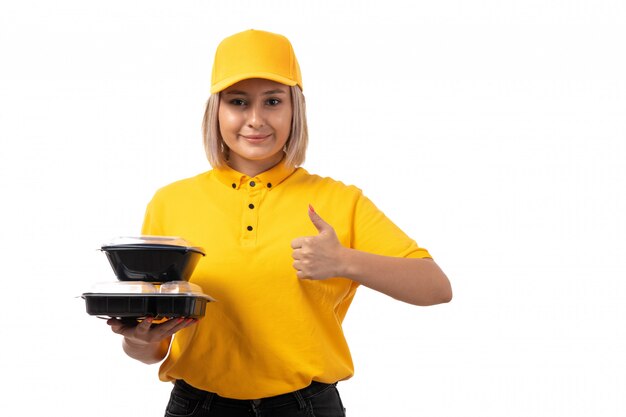 A front view female courier in yellow shirt yellow cap and black jeans holding bowls with food smiling on white